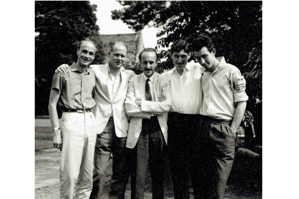 A group of men, smiling at the camera, wearing suits, shirts and trousers, with trees and an old brick building in the background.